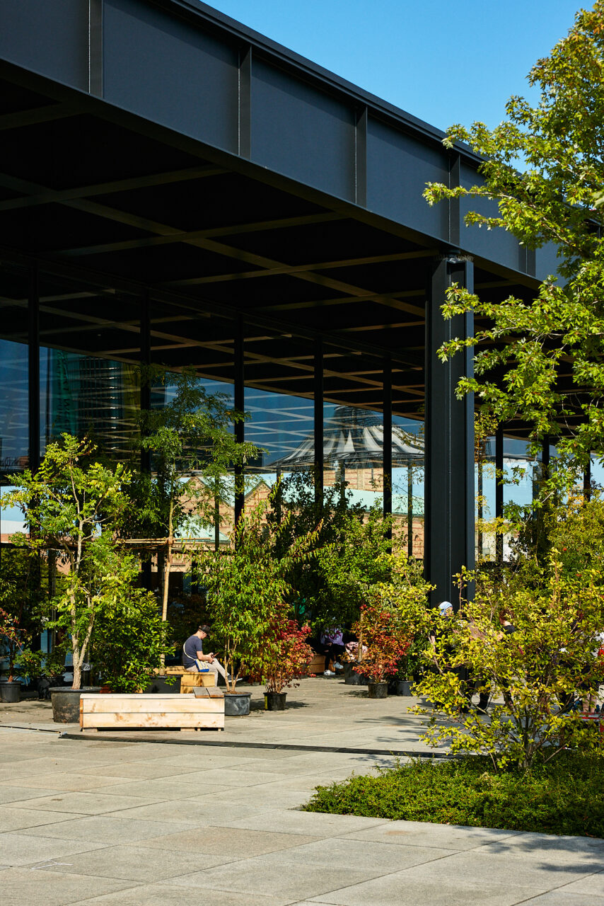 Moderne Architektur mit Glasfassade, umgeben von B&auml;umen und Pflanzen, im Freien sitzende Menschen unter blauem Himmel.