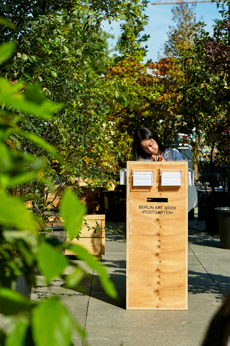 Teilnehmerin der Berlin Art Week schreibt an einem Holztisch, umgeben von Pflanzen und Kunstinstallationen.