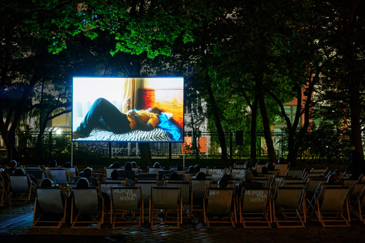 Open-Air-Kino mit Zuschauern auf Liegest&uuml;hlen, die einem Film auf einer gro&szlig;en Leinwand folgen, umgeben von B&auml;umen.