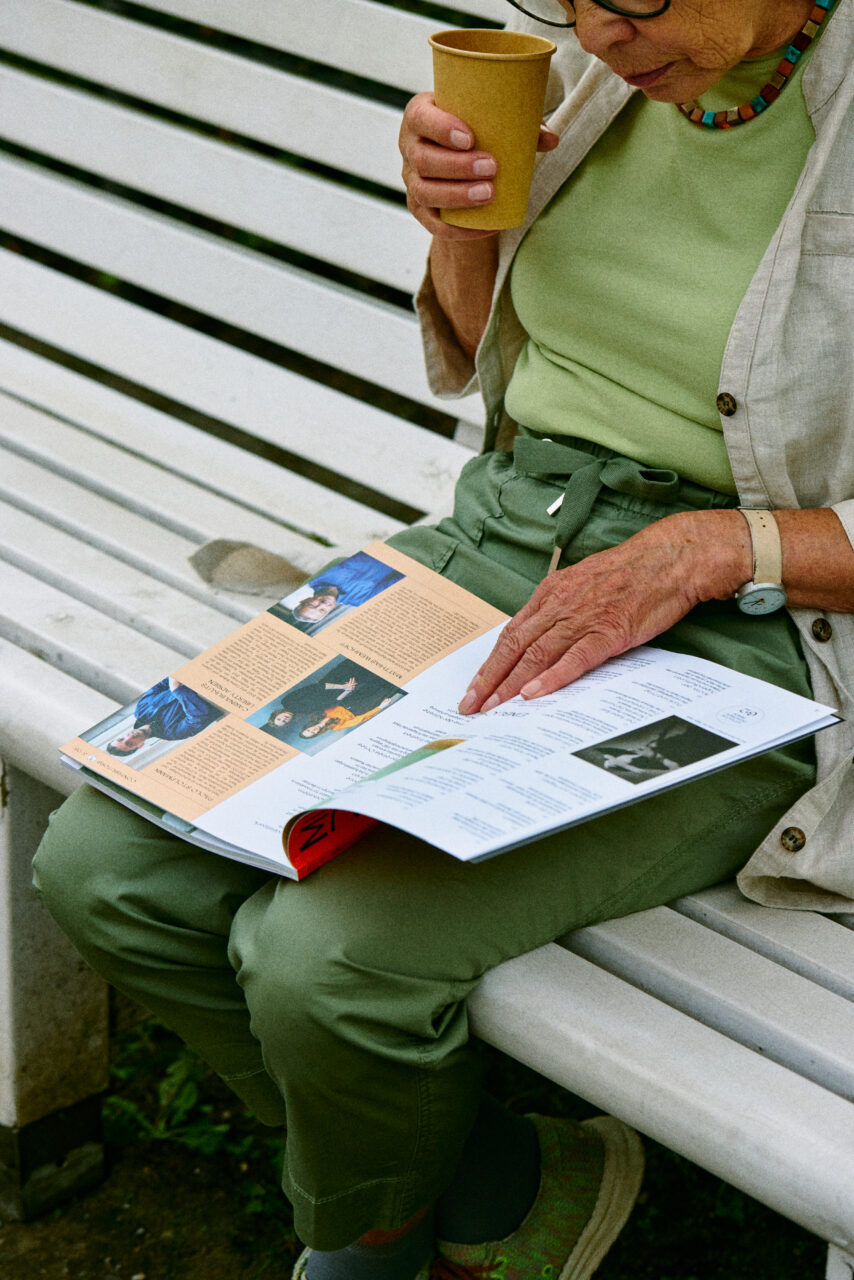 Eine Person sitzt auf einer Bank, h&auml;lt einen Becher und liest eine Zeitschrift mit bunten Bildern.